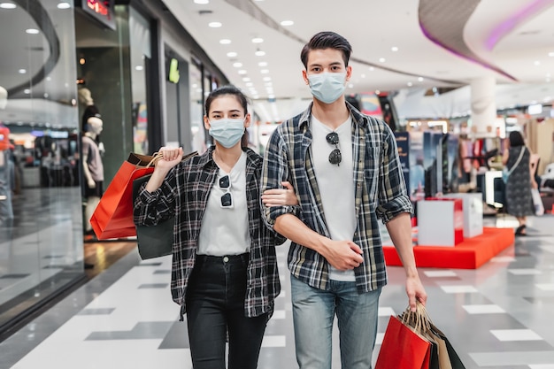 Young couple in protection mask holding multiple paper shopping bag walking in the corridor of large shopping mall