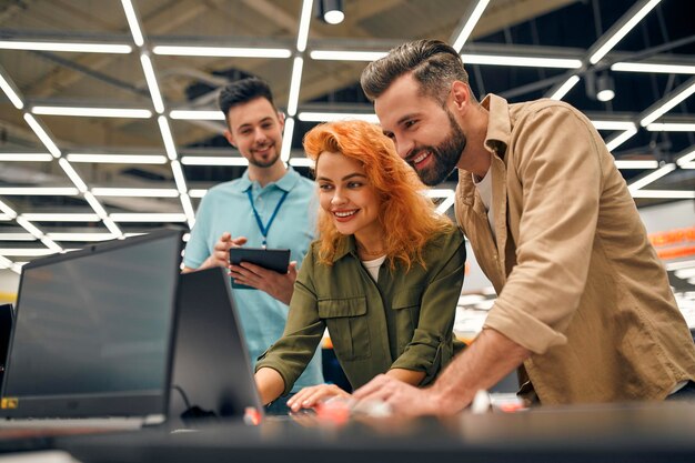 Young couple receives advice from seller in store of household
appliances and gadgets, buying new laptop. male consultant helps in
choosing laptop.