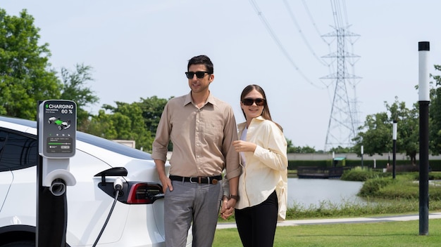 Young couple recharge EV car battery at charging station connected to power grid tower electrical industrial facility as electrical industry for eco friendly vehicle utilization Expedient