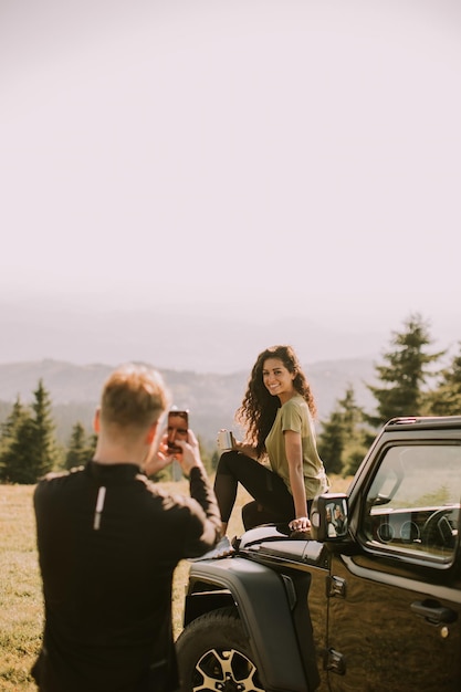 Young couple relaxing on a terrain vehicle hood and taking photo with mobile phone at countryside