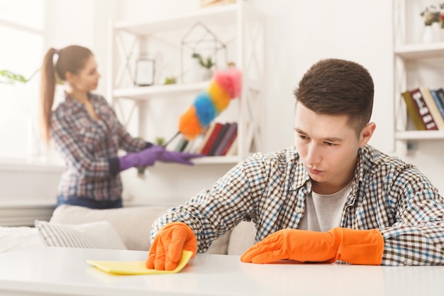 Young couple in rubber gloves cleaning home, wiping dust with towel and feather brush in living-room, copy space, selective focus. Housekeeping and cleaning service concept