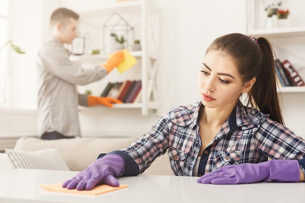 Young couple in rubber gloves cleaning home, wiping dust with towel in living-room, copy space, selective focus. Housekeeping and cleaning service concept