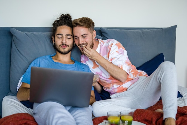 ベッドで朝食をとっている同性の若いカップル、ラップトップで映画を見ている男性のゲイのカップル、封鎖でのビデオ通話、Covid19の流行による検疫