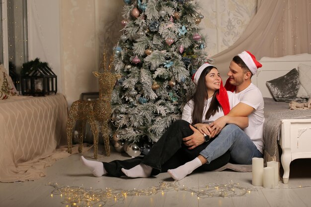 young couple in Santa Claus hats is sitting hugging on the floor by the bed and the Christmas tree