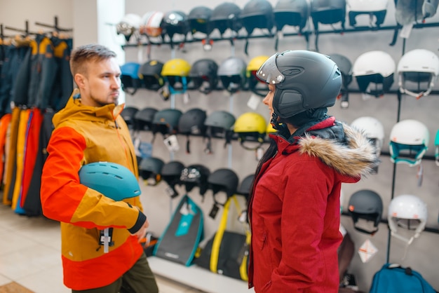 Giovani coppie alla vetrina che provano sui caschi per sci o snowboard, vista laterale, negozio di articoli sportivi.