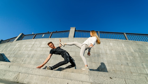 Young couple showing motions near the grey rock wall.Flying hair. Motion picture. Woman knocks a man with a leg. Stylish photo. Blue sky above.