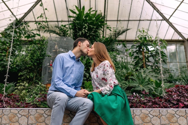 Young couple sits on a bench and kisses in a green botanical garden