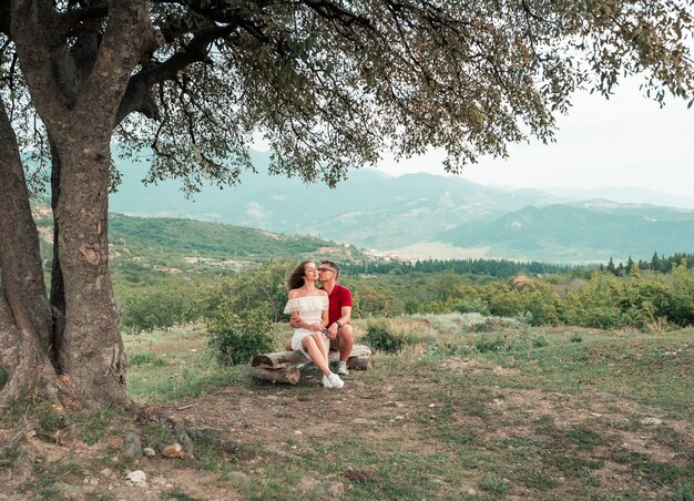 Young couple sits on a bench near a beautiful tree on mountains