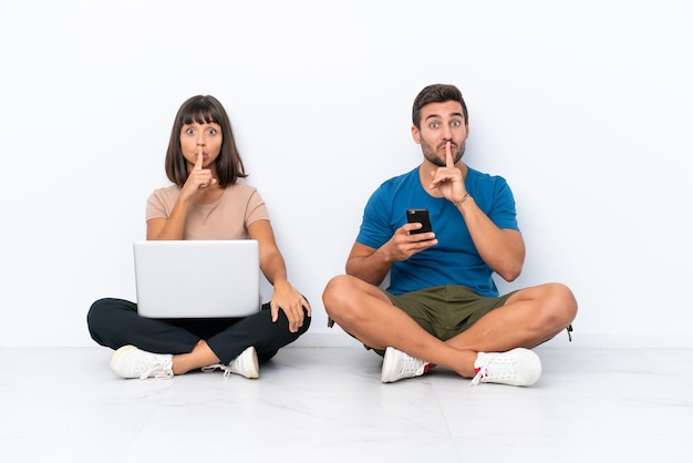 Young couple sitting on the floor holding pc and mobile phone isolated on white background showing a sign of silence gesture putting finger in mouth