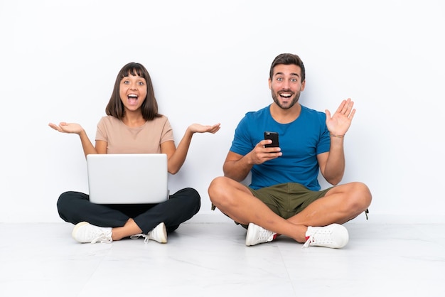 Молодая пара сидит на полу, держа компьютер и мобильный телефон на белом фоне с удивленным и шокированным выражением лица