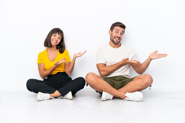 Young couple sitting on the floor isolated on white background extending hands to the side for inviting to come