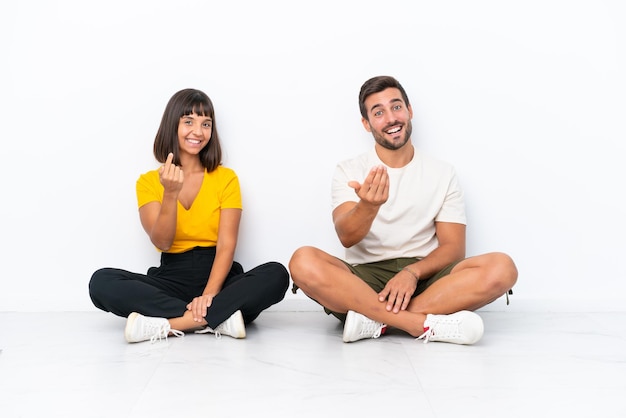 Young couple sitting on the floor isolated on white background inviting to come with hand. Happy that you came