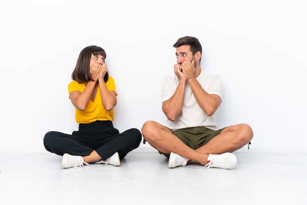 Young couple sitting on the floor isolated on white background is a little bit nervous and scared putting hands to mouth