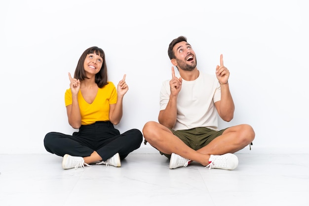 Photo young couple sitting on the floor isolated on white background pointing with the index finger a great idea