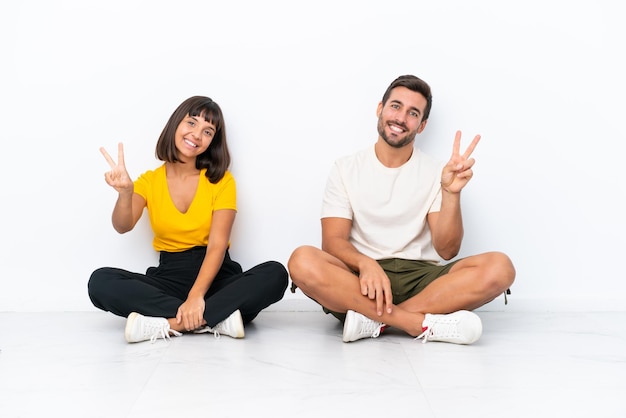 Young couple sitting on the floor isolated on white background smiling and showing victory sign