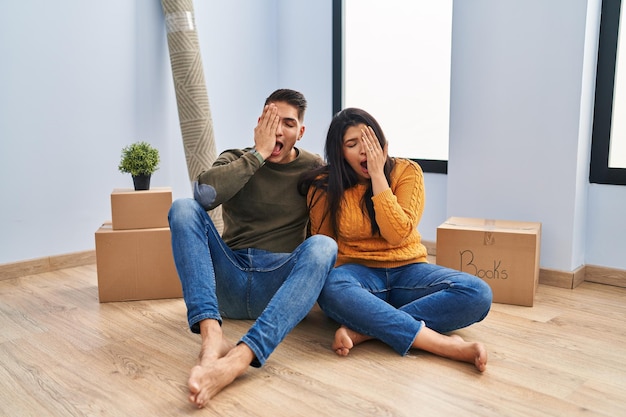 Young couple sitting on the floor at new home yawning tired covering half face, eye and mouth with hand. face hurts in pain.