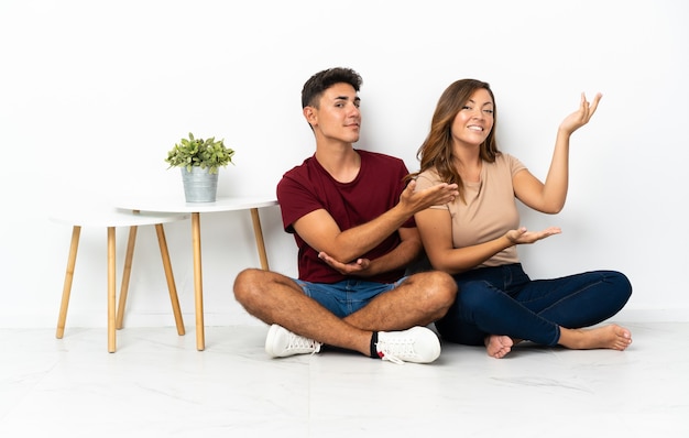 Young couple sitting on the floor on white extending hands to the side for inviting to come