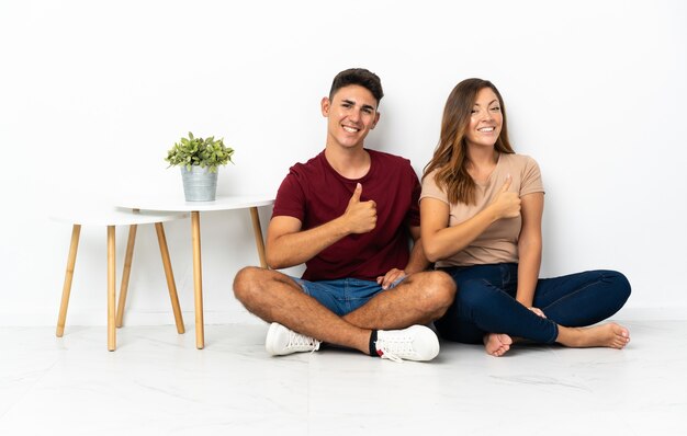 Young couple sitting on the floor on white giving a thumbs up gesture