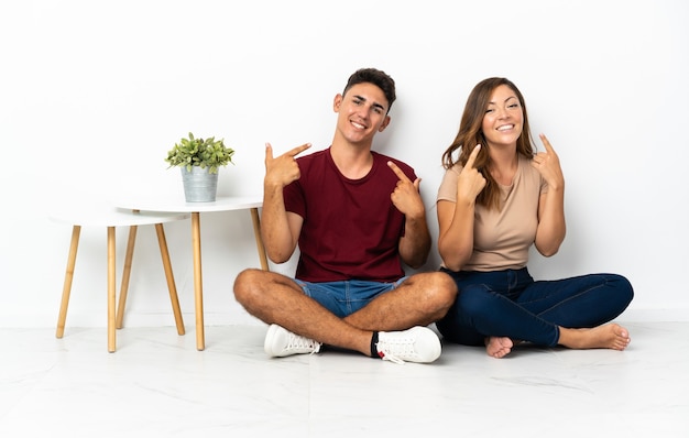 Young couple sitting on the floor on white giving a thumbs up gesture