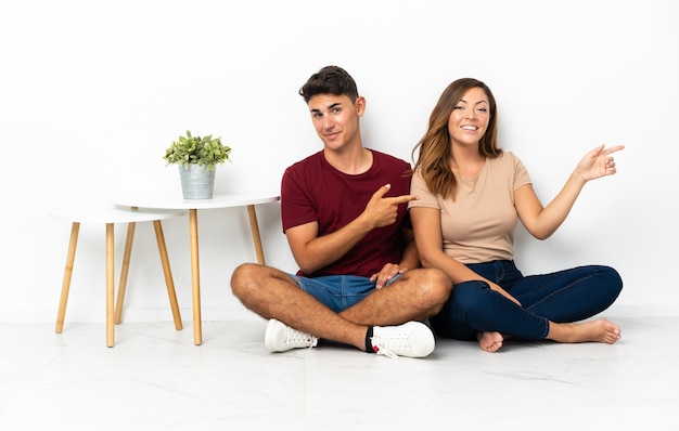 Photo young couple sitting on the floor on white pointing finger to the side