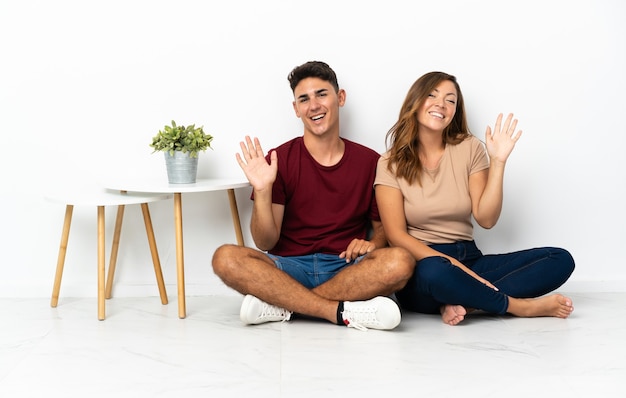 Photo young couple sitting on the floor on white saluting with hand with happy expression