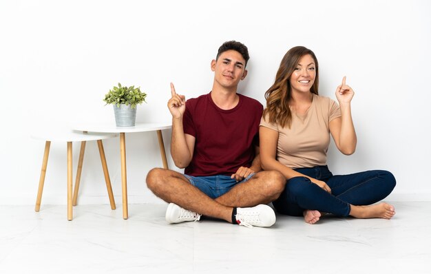 Photo young couple sitting on the floor on white showing and lifting a finger in sign of the best