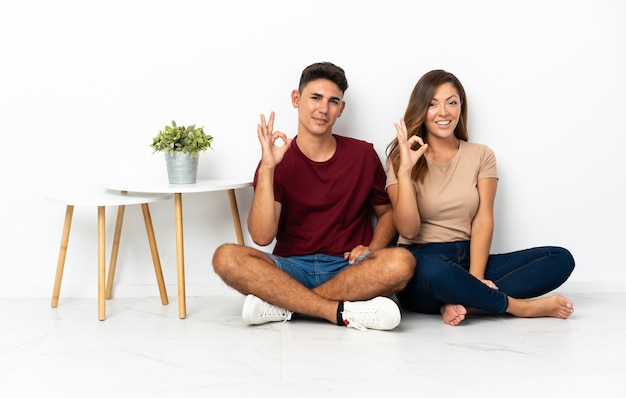 Photo young couple sitting on the floor on white showing ok sign with fingers