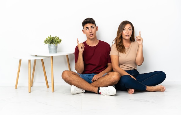 Young couple sitting on the floor on white thinking an idea pointing the finger up