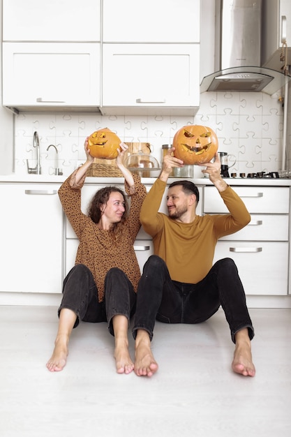 Photo young couple sitting on the kitchen floor and holding halloween pumpkins