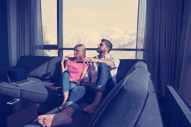 Young couple sitting on sofa near the window watching television together at luxury home