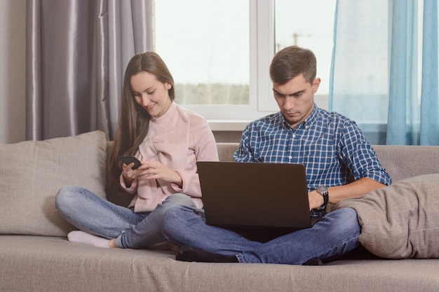 young couple sitting on the sofa in the room, busy with their gadgets, communication problems of modern society