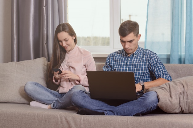 Young couple sitting on the sofa in the room, busy with their gadgets, communication problems of modern society