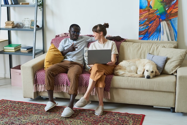 Young couple sitting on sofa with laptop and talking to each other in the room at home