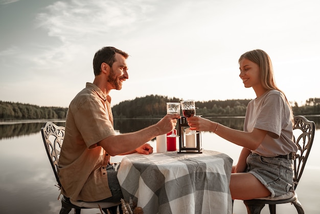 記念日を祝って、手をつないで森の湖で一緒にワインを飲みながらテーブルに座っている若いカップル。愛は空中にあり、ラブストーリーのコンセプトです。キャンドルとロマンチックなデート。外で2人分の夕食