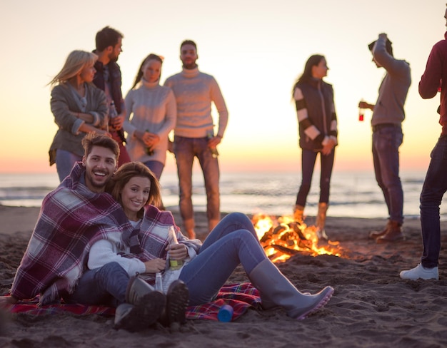 Young couple sitting with friends around campfire on the beach\
at sunset drinking beer