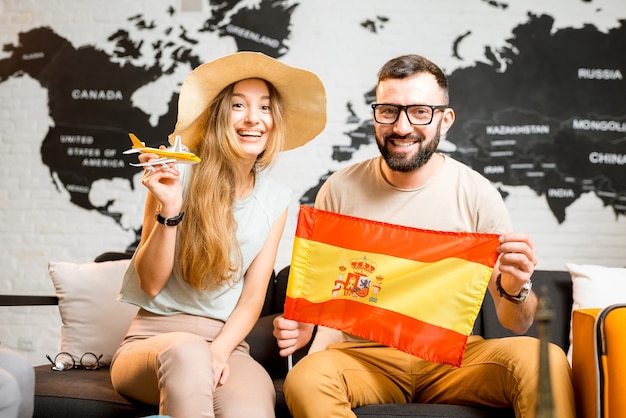 スペインへの旅行の準備をしている世界地図の背景の旅行代理店のオフィスでスペイン国旗と一緒に座っている若いカップル