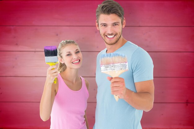 Young couple smiling and holding paintbrushes against wooden planks background