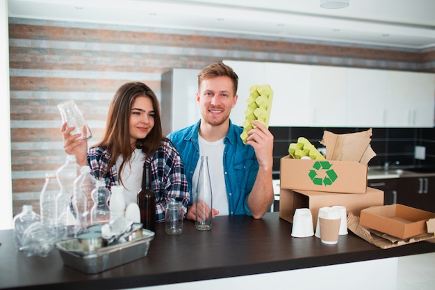 A young couple sorts trash in the kitchen. Young man and woman are sorting recyclables in the kitchen. There is cardboard, paper, iron, plastic and glass and other materials that can be recycled.