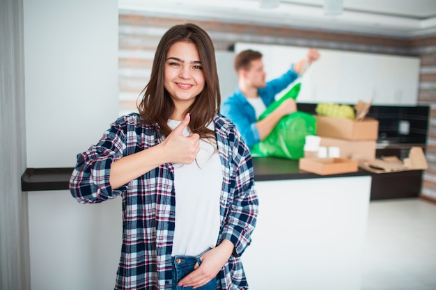 A young couple sorts trash in the kitchen. Young man and woman are sorting recyclables. Young woman showing thumbs up.