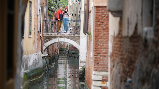 A young couple standing on the bridge between two buildings and taking photo Venice Italy