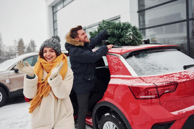 Young couple standing near car with tree on the top. Together outdoors at winter time.