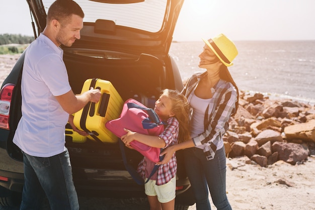 Giovani coppie che stanno vicino allo stivale aperto dell'automobile con le valigie e le borse. papà, mamma e figlia viaggiano in riva al mare o sull'oceano o sul fiume. giro estivo in automobile