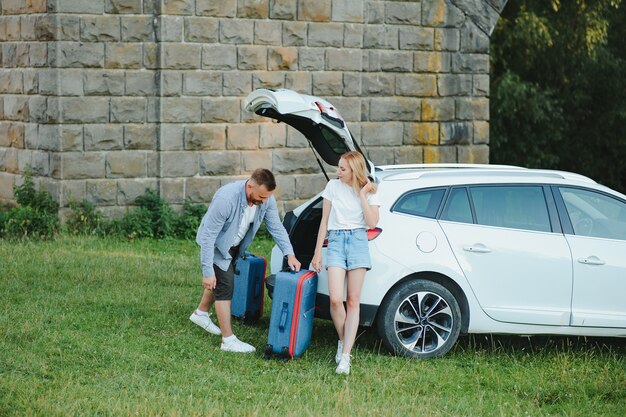 スーツケースを持って開いた車のトランクの近くに立って、カメラを見て、屋外で若いカップル