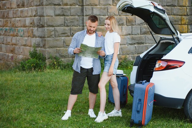 Young couple standing near the opened car boot with suitcases, looking to the camera, outdoors