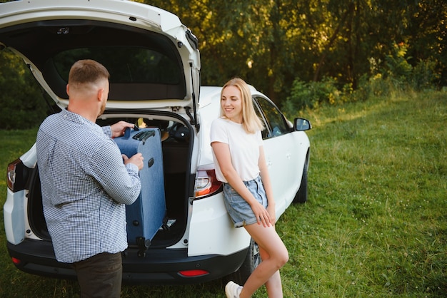 Young couple standing near the opened car boot with suitcases, looking to the camera, outdoors