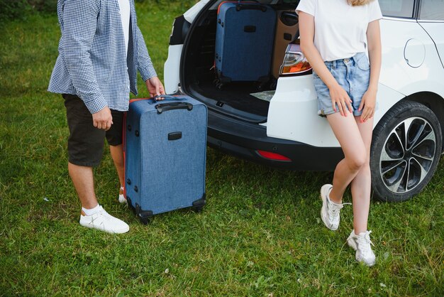 スーツケースを持って開いた車のトランクの近くに立って、カメラを見て、屋外で若いカップル