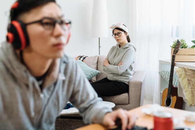 젊은 부부는 여름날 집에 있다. 헤드폰을 끼고 컴퓨터로 온라인 게임을 하는 아시아 남성의 시야가 흐릿한 반면 좌절한 여자친구는 소파에 앉아 실내에서 남자친구를 바라보고 있습니다.