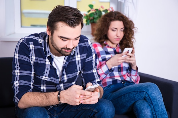 Young couple surfing on their cell phones sitting on sofa at home