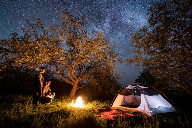 Young couple tourists standing at a campfire near tent under trees and beautiful night sky full of stars and milky way. Night camping