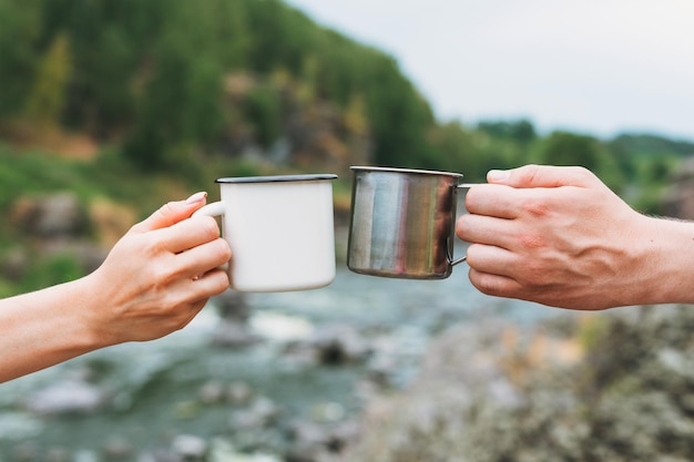 Young couple travelers holding metal mugs on mountain river background Local tourism weekend trip